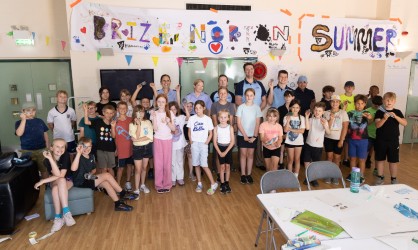 An image showing a large group of attending children and adults posing together indoors under a colourful banner that reads "BRIZE NORTON SUMMER," with everyone standing on a light wood floor next to craft tables.